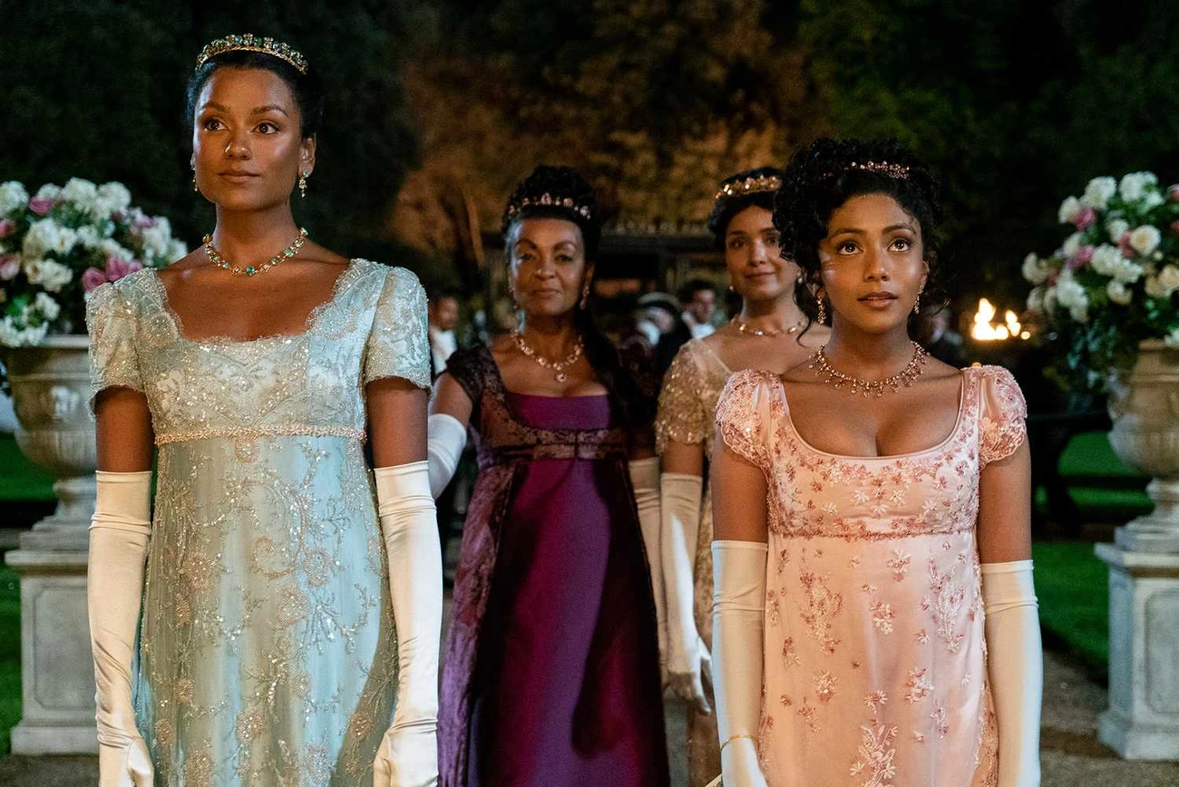 Four women in elaborate Regency-era dresses and gloves stand outdoors at night amid floral arrangements and soft lighting.