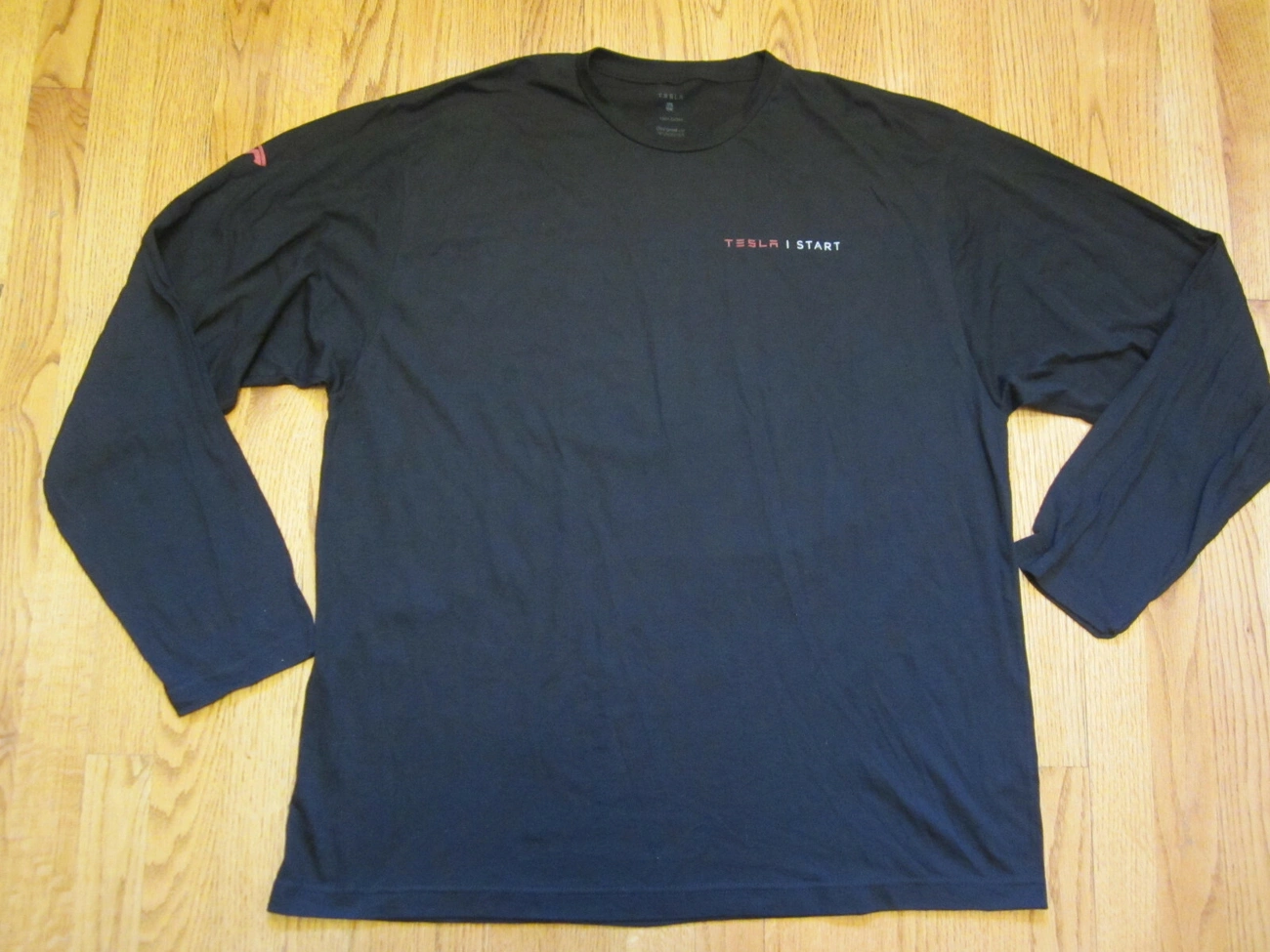 Black long-sleeve Tesla START student automotive tech mechanic T-shirt laid flat on a wooden floor, size 2XL.